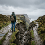 Person wearing green jacket and backpack standing near a 'Path Closed' sign on a cliffside trail overlooking the ocean